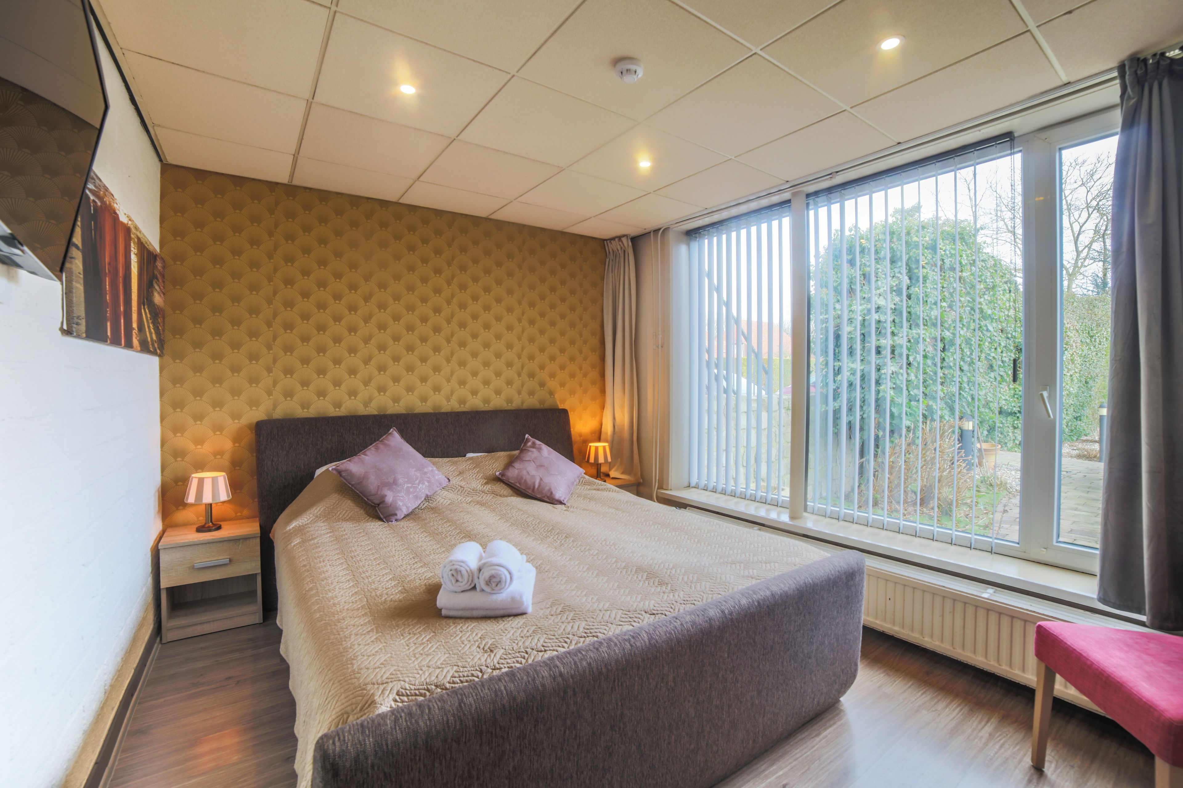Een hotelkamer met een groot bed, twee bedlampen, een raam met zonwering en uitzicht op groen.