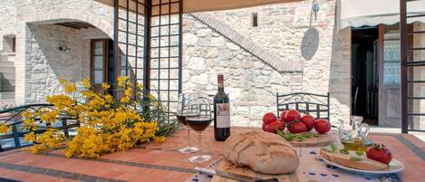 An outdoor dining area with a table set for two, featuring a loaf of bread, tomatoes, a bottle of wine, and a vase of yellow flowers.