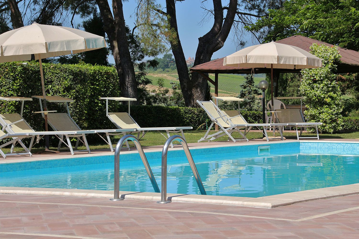 Ein Schwimmbecken mit Liegestühlen und Sonnenschirmen, umgeben von Bäumen und blauem Himmel.