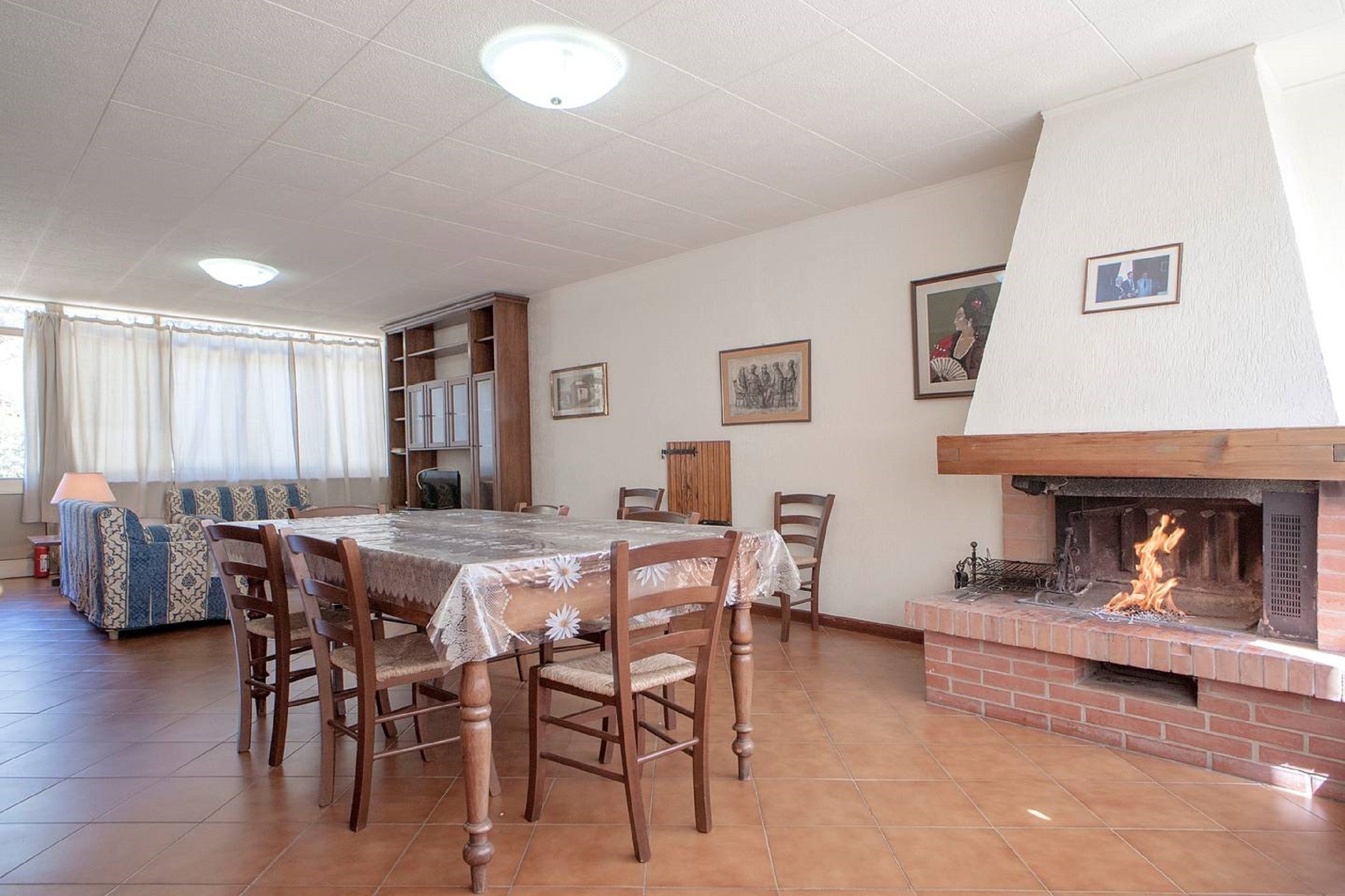 A dining area with a brick fireplace, wooden chairs, a table with a lace tablecloth, and a sofa with patterned cushions.