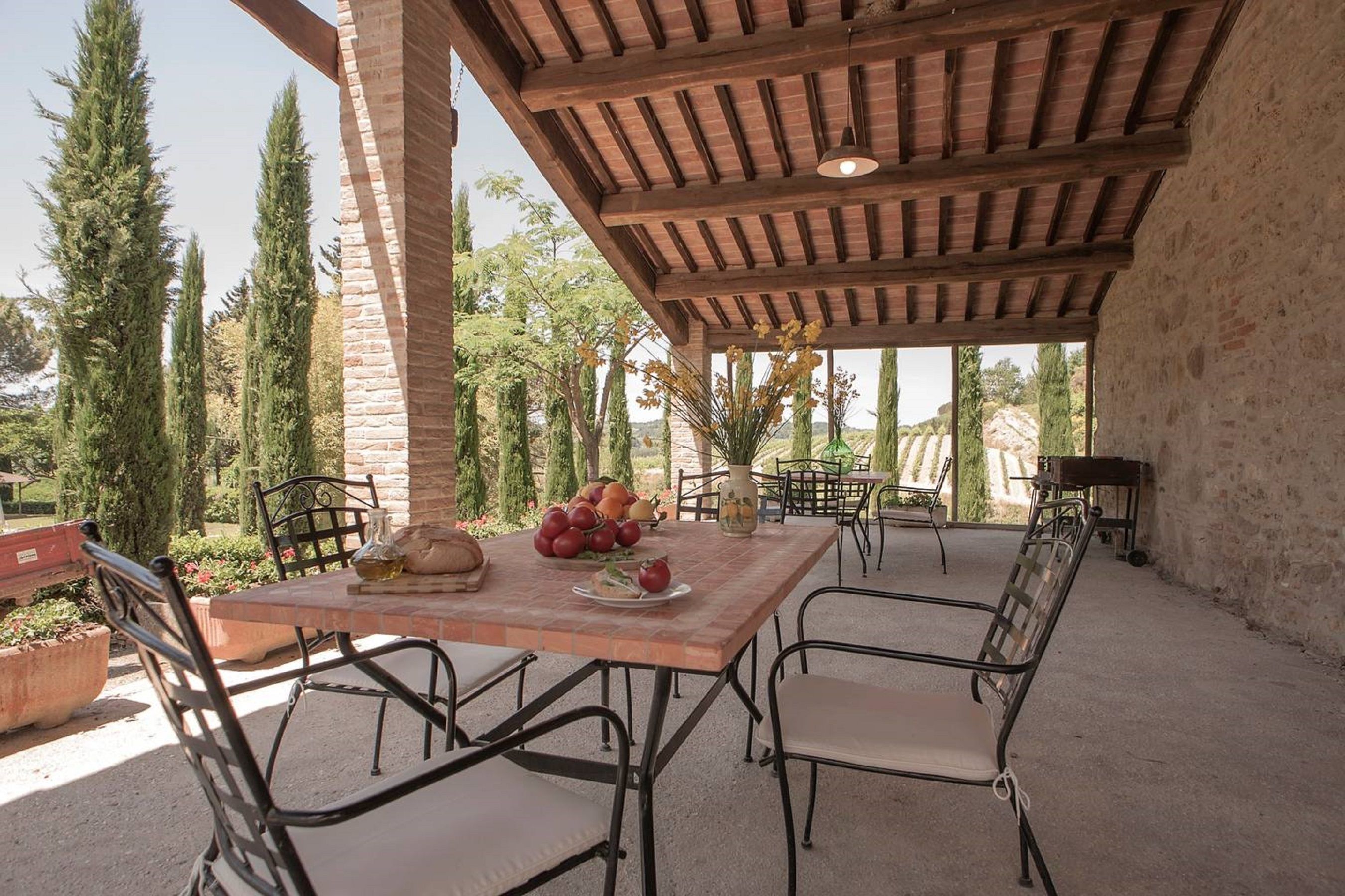 Eine Terrasse im Freien mit Holztisch und Metallstühlen, umgeben von hohen Bäumen und einer Steinmauer.