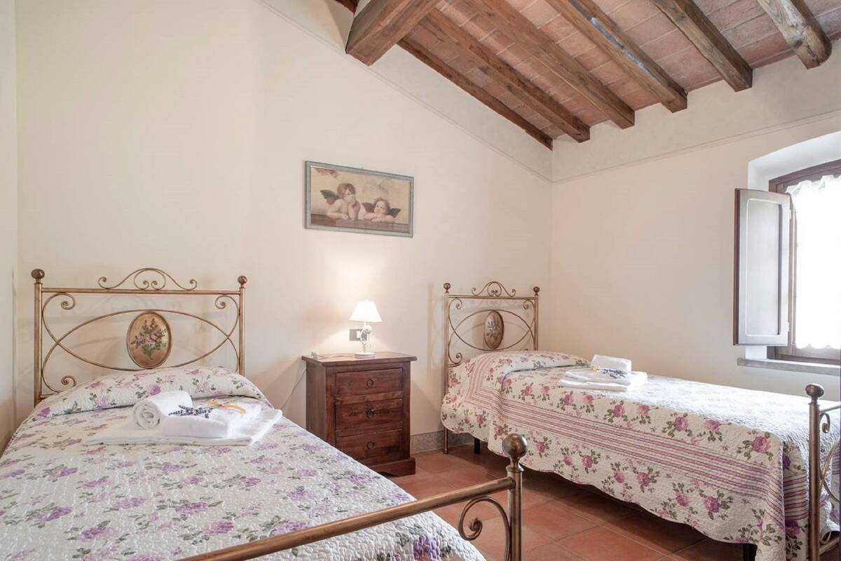 Une chambre avec deux lits, des têtes de lit en bois, une table de chevet avec une lampe, un tableau au mur et une fenêtre avec des rideaux.