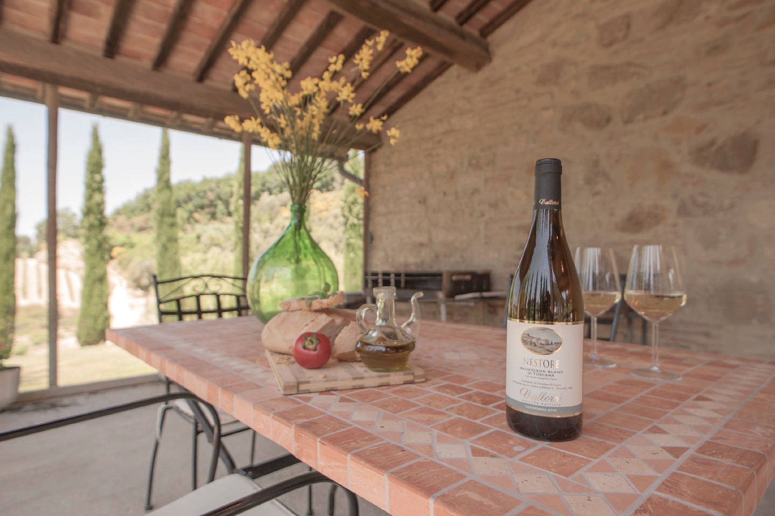 A table with a bottle of wine, glasses, a loaf of bread, a tomato, and a vase with flowers.