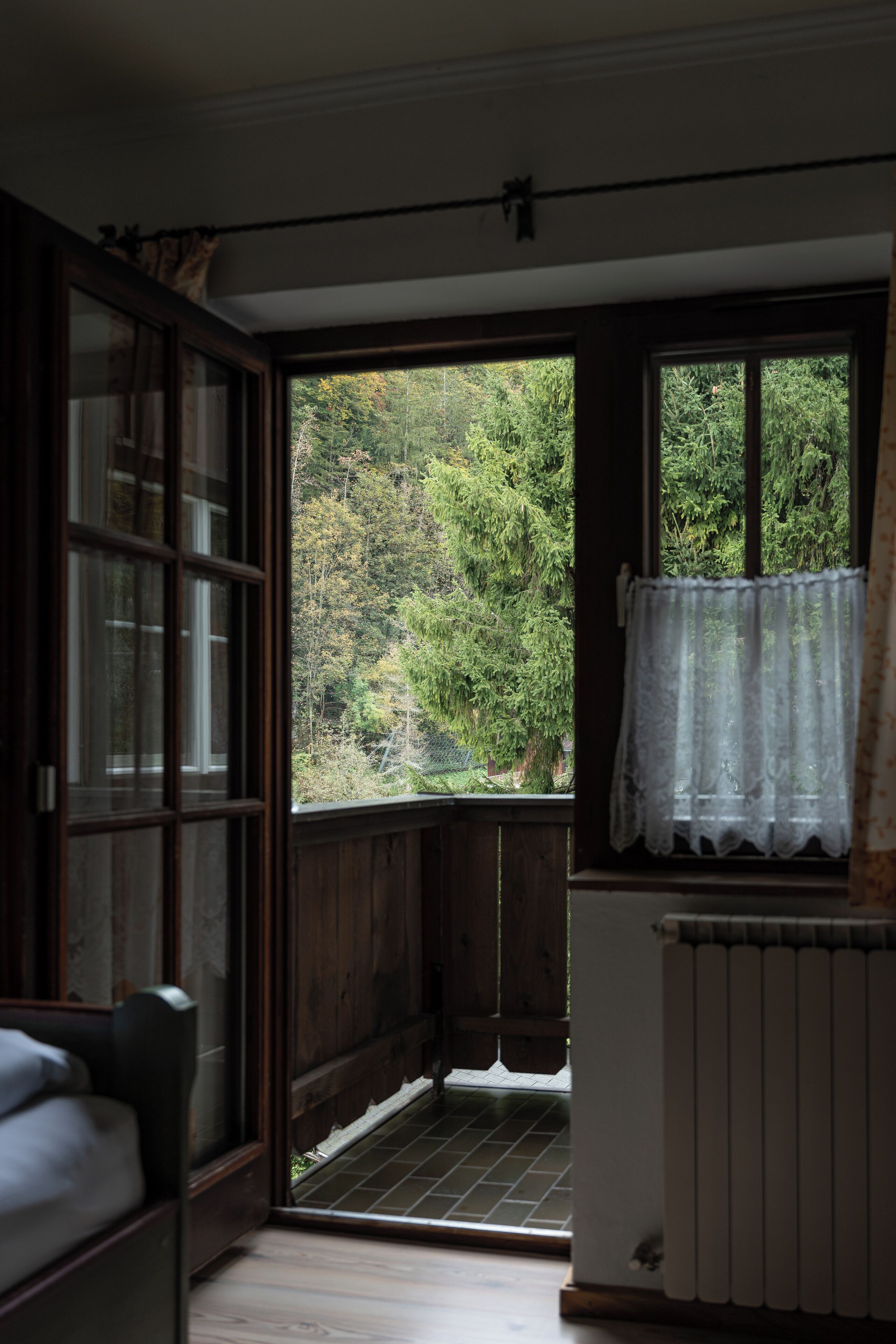 Une pièce avec un plancher en bois, un radiateur, une fenêtre ornée d’un rideau en dentelle et une vue sur une forêt.