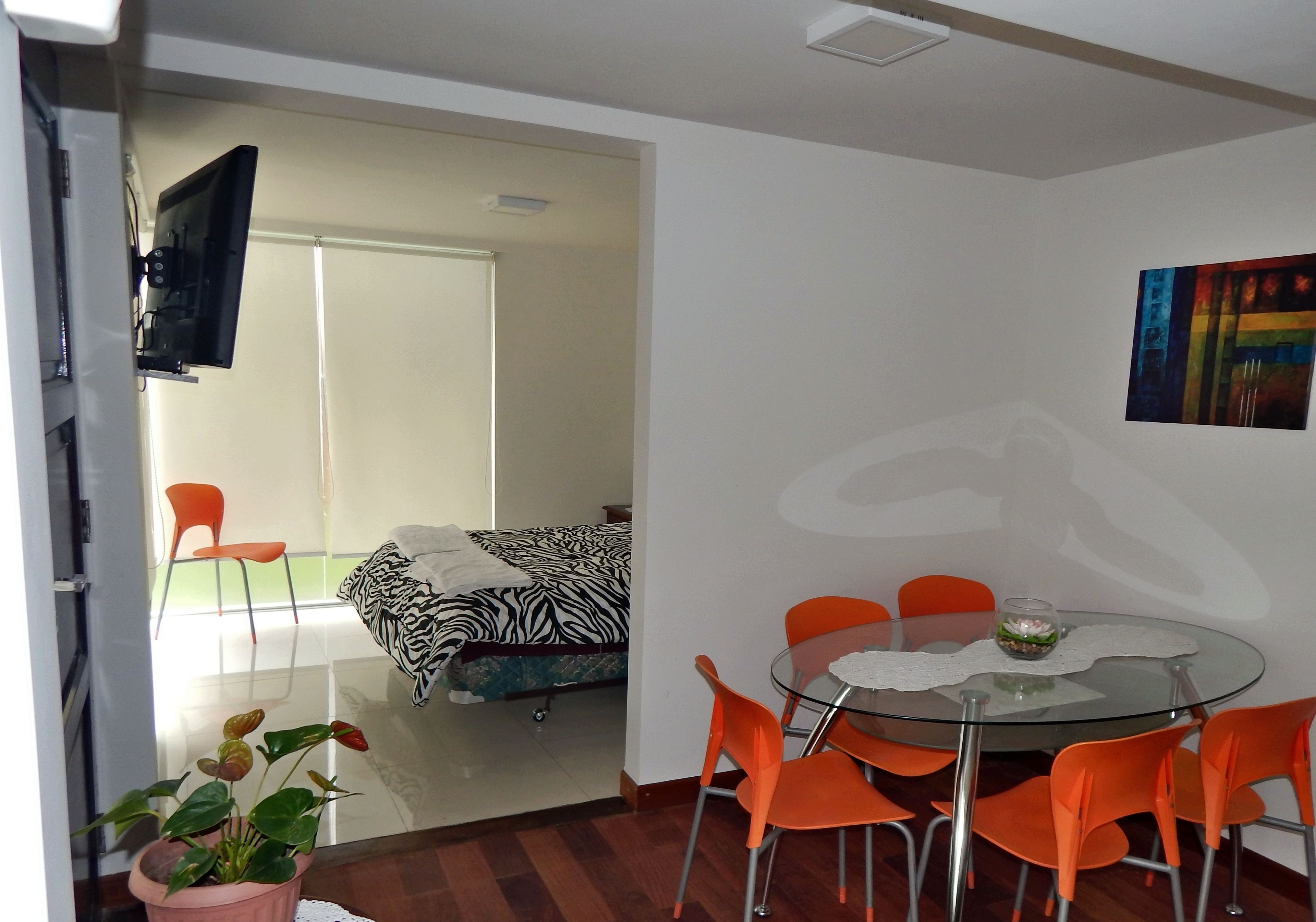 A modern living room with a glass dining table, orange chairs, a zebra-patterned sofa, a flat-screen TV, and a potted plant.