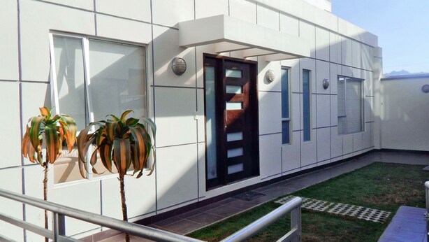 Un edificio moderno con fachada blanca y limpia, una puerta central y un pequeño espacio con césped y plantas en macetas.