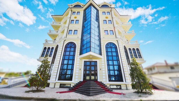 A large, multi-story building with a central glass tower, a bar at the entrance, and a symmetrical facade with multiple windows and balconies. - Hotel Luani Arte (Shkodër)