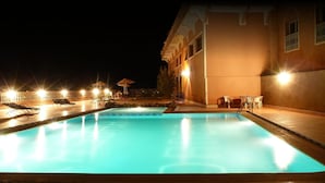 A nighttime swimming pool area with lounge chairs and umbrellas, surrounded by a building with a tiled roof and a brick wall. - La Kasbah De Dades (Boumalne Dades)
