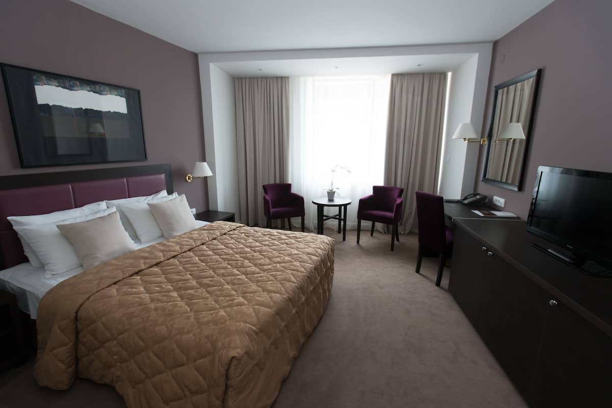 Ein Hotelzimmer mit einem großen Bett, einem Schreibtisch mit Fernseher, zwei Sesseln, einem kleinen Tisch mit Pflanze und einem Fenster mit Vorhängen.