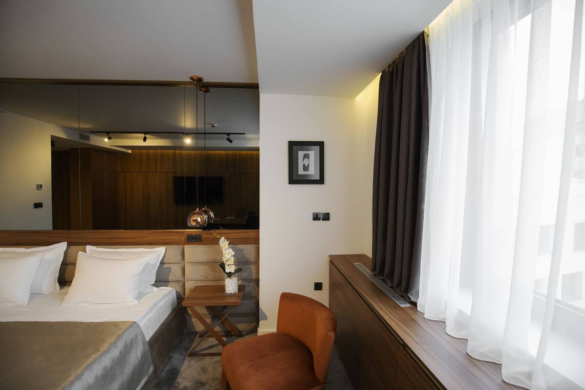 Ein Hotelzimmer mit einem Bett, einem kleinen Tisch mit Blumen, einem Sessel, einem Fenster mit Vorhängen und einer Küchenzeile im Hintergrund.