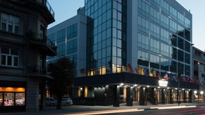 Een modern hotel met een glazen gevel, een opvallend 'HOTEL'-bord en een klassiek pand links.