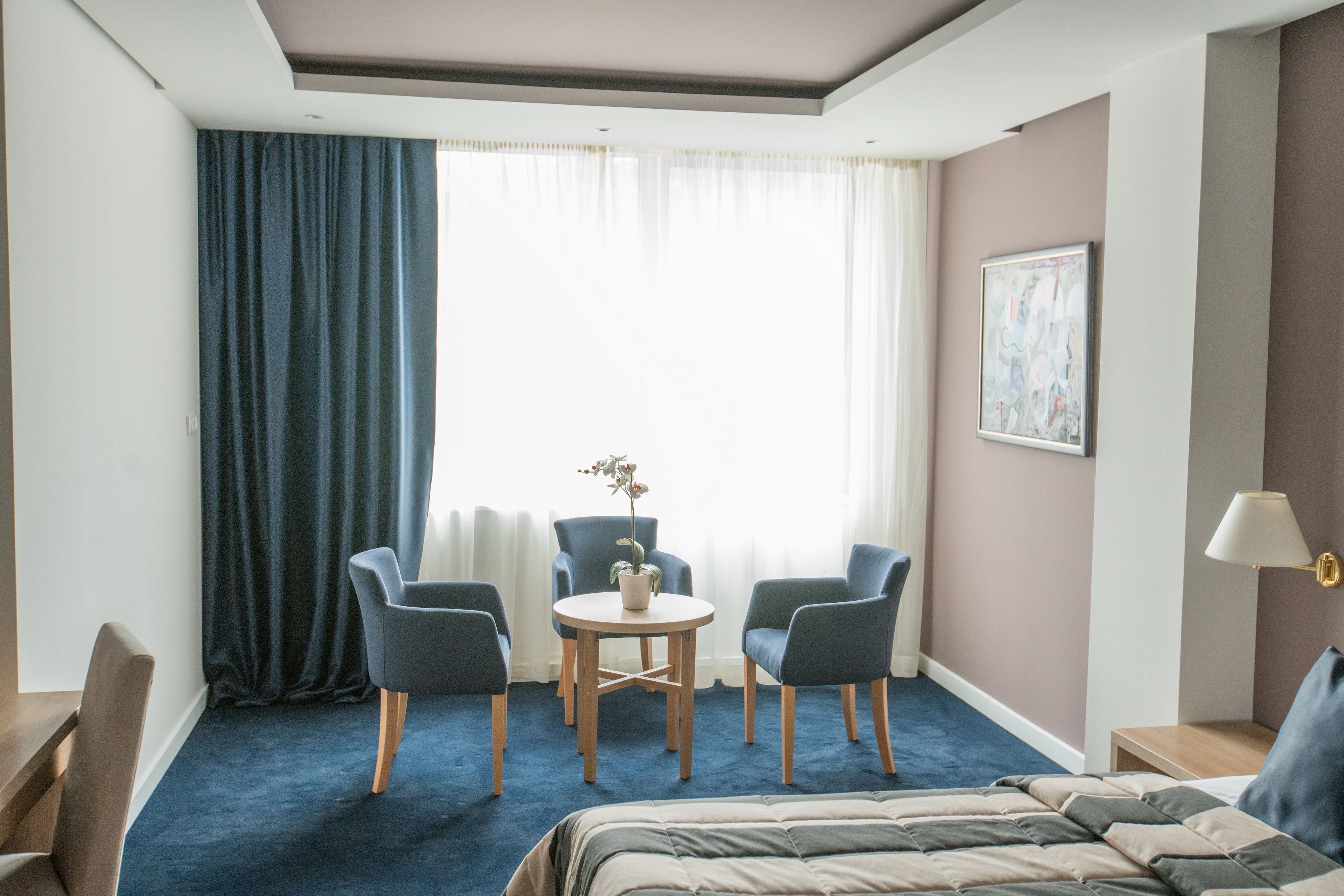 A hotel room with a bed, two armchairs, a small table with a plant, a painting on the wall, and a window with curtains.