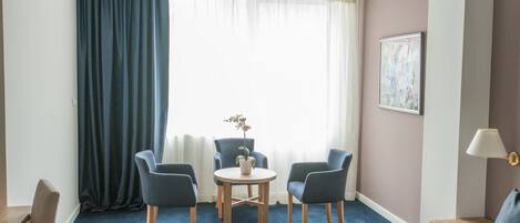Una habitación de hotel con una cama, dos sillones, una mesita con una planta, un cuadro en la pared y una ventana con cortinas.