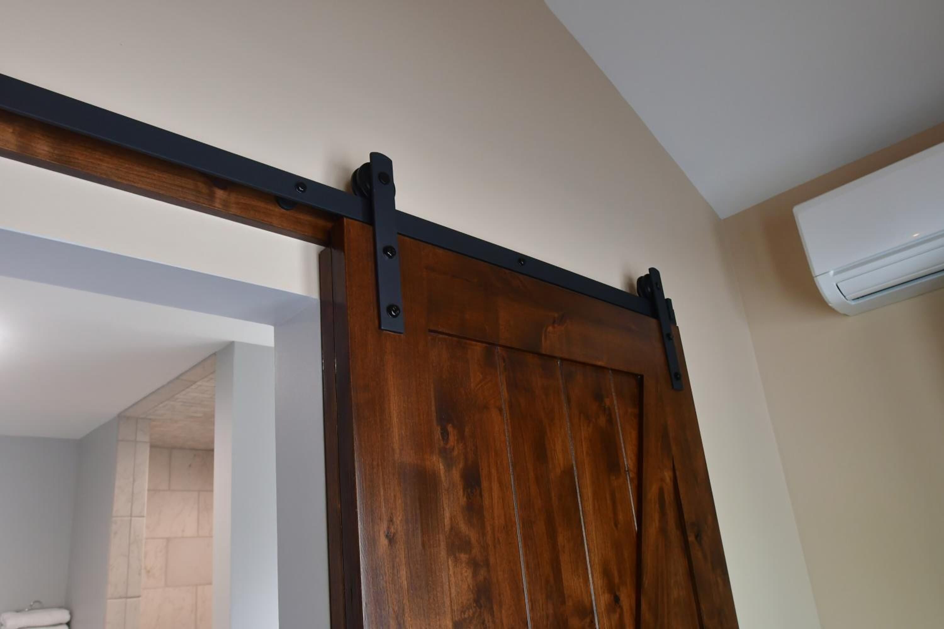 Una puerta corrediza de madera, con riel de metal negro, en una habitación con paredes color beige y un equipo de aire acondicionado en la pared.