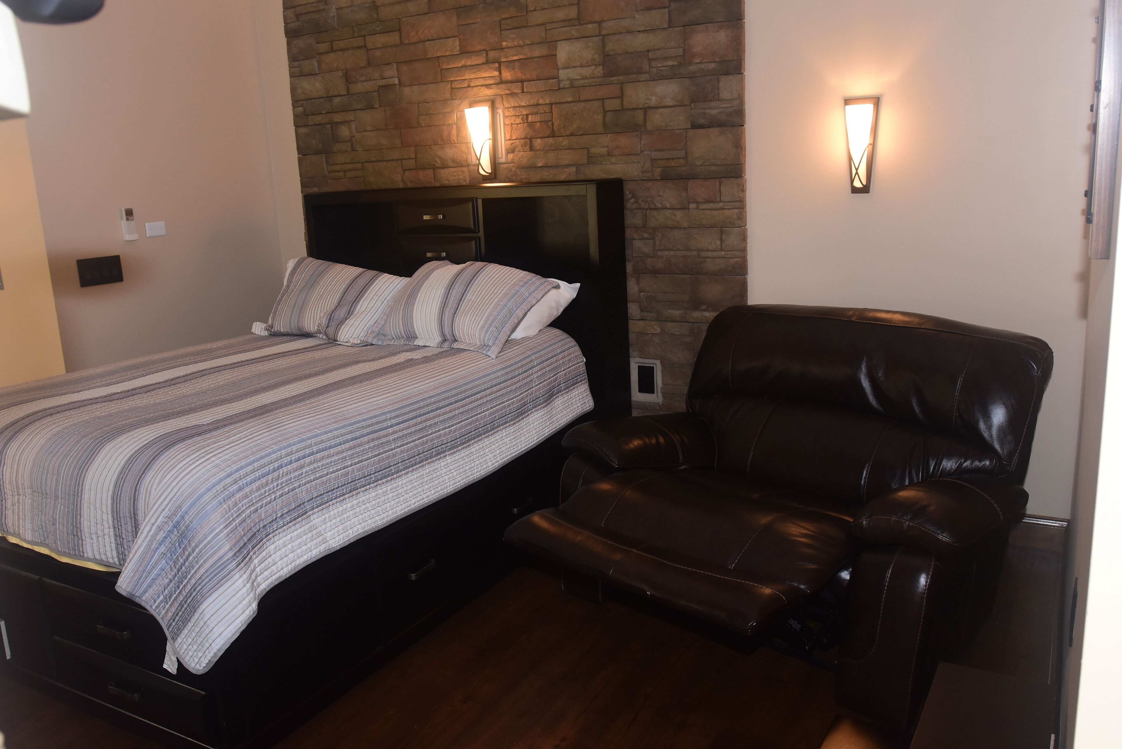 A bedroom with a bed, a leather chair, and two wall-mounted lights.