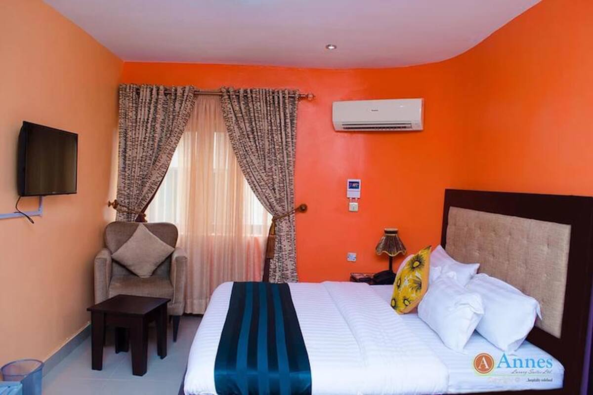A hotel room with a bed, a chair, a table, an air conditioning unit, and a television.