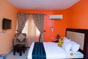 A hotel room with a bed, a chair, a table, an air conditioning unit, and a television. - Annes Luxury Suites Ltd (Lagos)