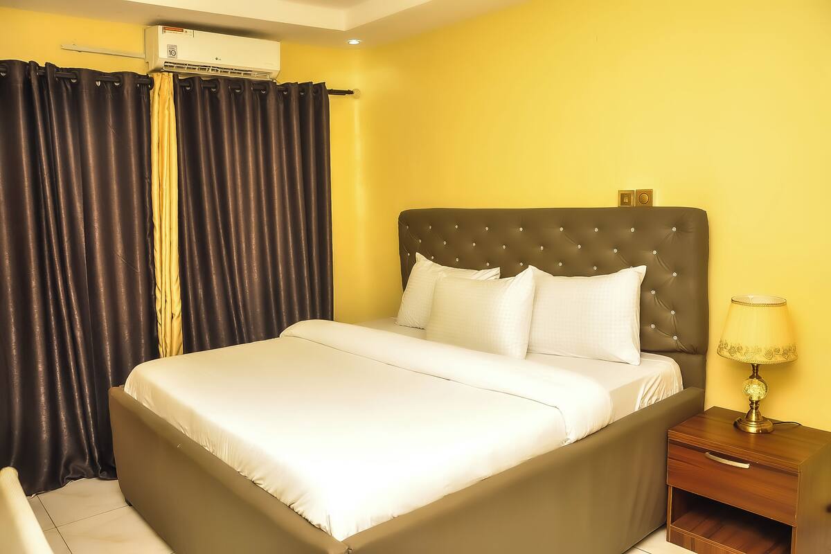 A neatly made bed with a headboard, a bedside table with a lamp, and a window with curtains.