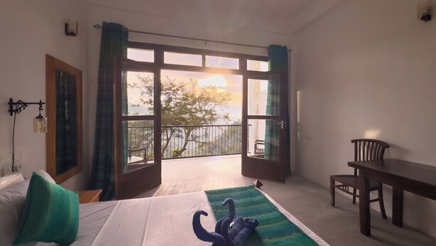 A hotel room with a bed, a desk, and a balcony with a view of trees and the sky. - Ella Alpine Resort (Ella)