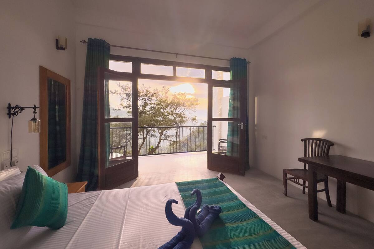 A hotel room with a bed, a desk, and a balcony with a view of trees and the sky.