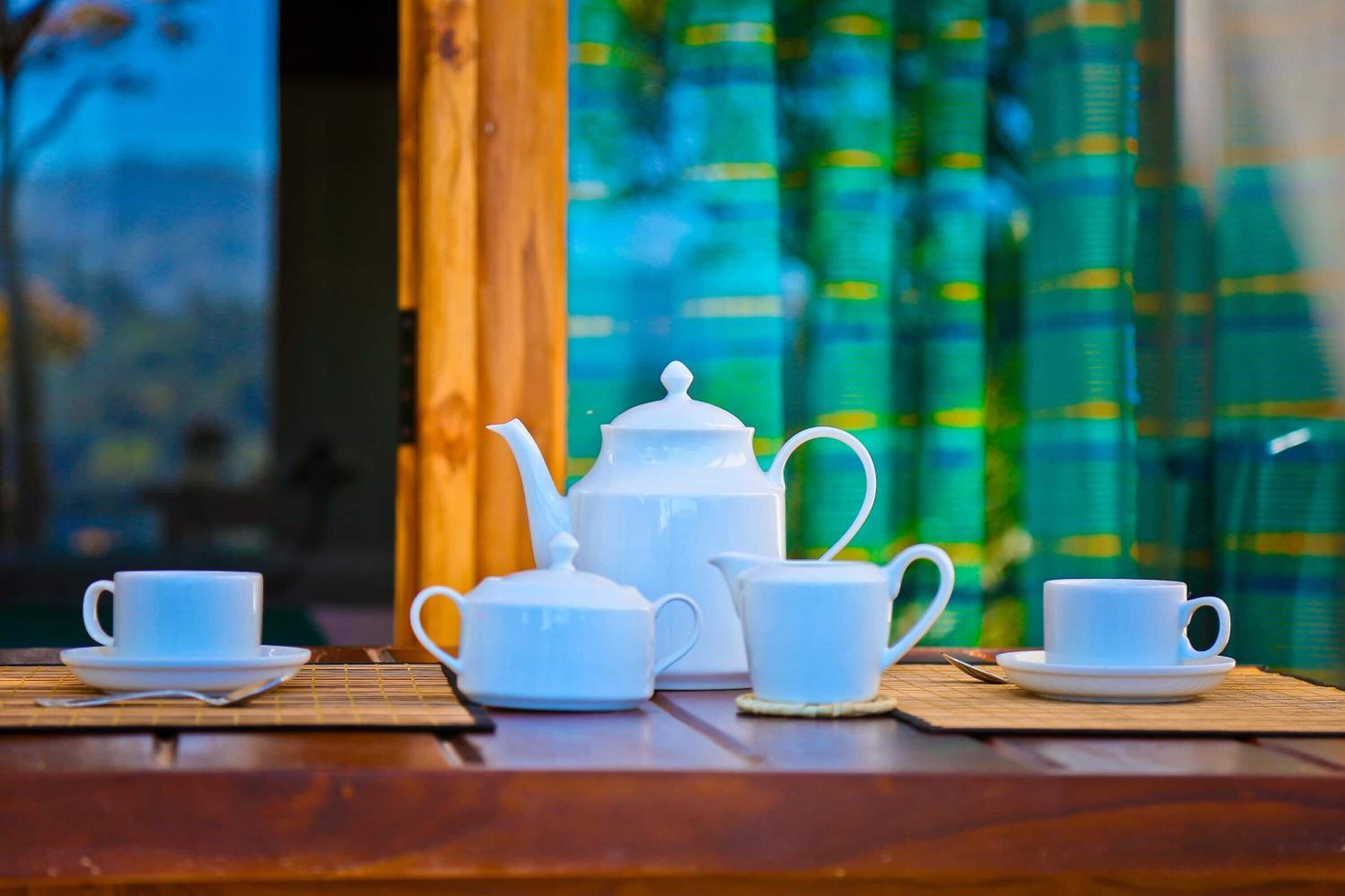 A tea set on a wooden table with a view of green curtains and a window in the background.