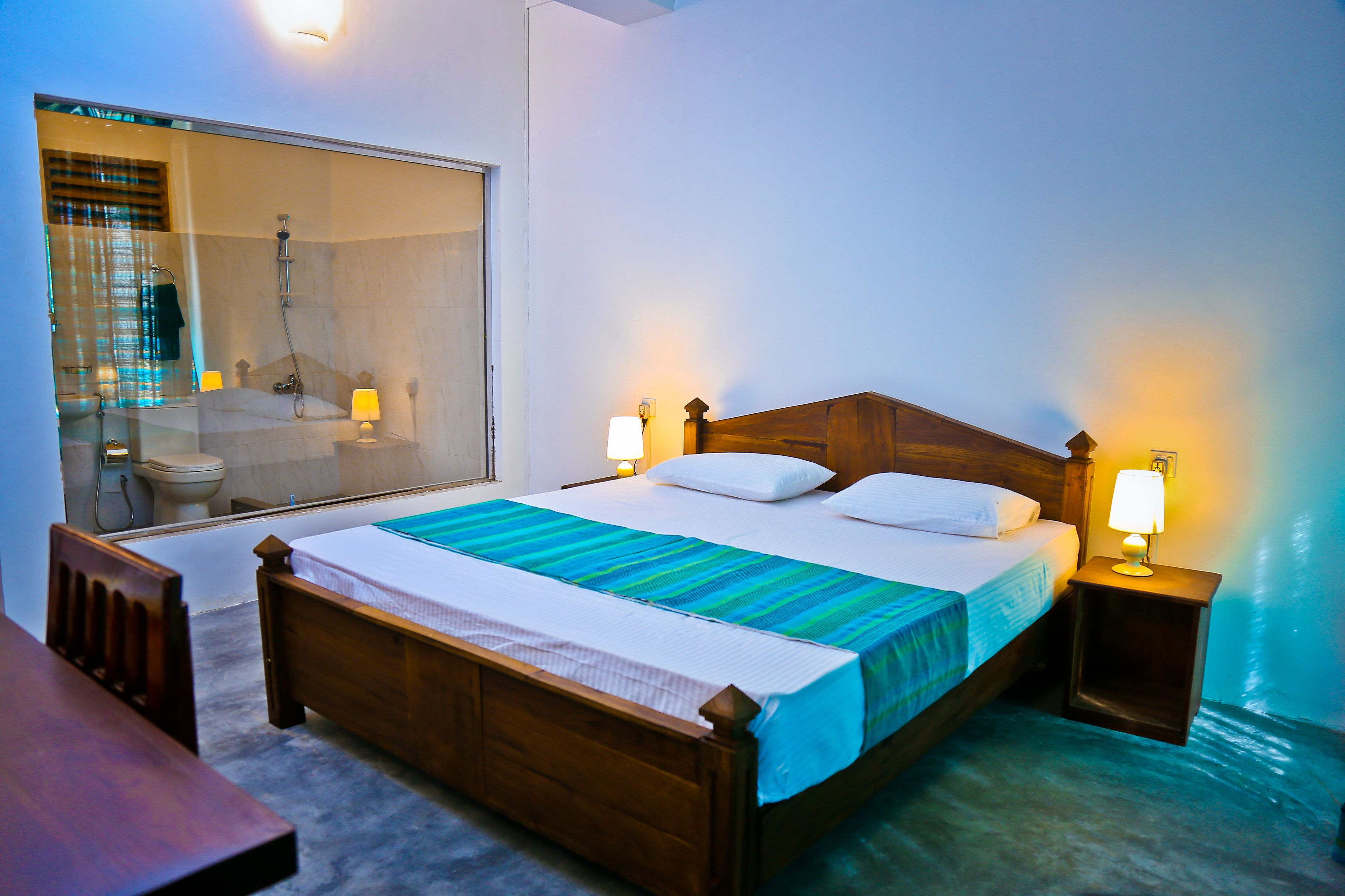 A neatly made bed with a wooden headboard, two bedside lamps, and a large mirror reflecting the bathroom.