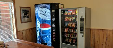 A room with a wooden table, a Pepsi vending machine, and a snack vending machine.