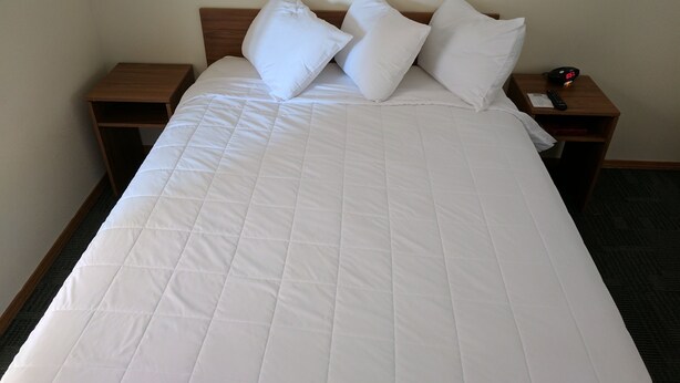 A neatly made bed with a white quilt and pillows, a wooden headboard, and two nightstands with a clock and a remote control. - Travelodge by Wyndham Three Forks (Three Forks)