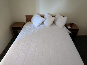 A neatly made bed with a white quilt and pillows, a wooden headboard, and two nightstands with a clock and a remote control. - Travelodge by Wyndham Three Forks (Three Forks)