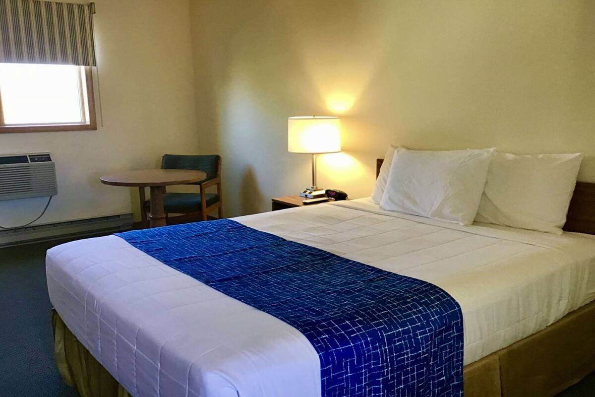 A hotel room with a bed, a chair, a table, a lamp, and an air conditioning unit.
