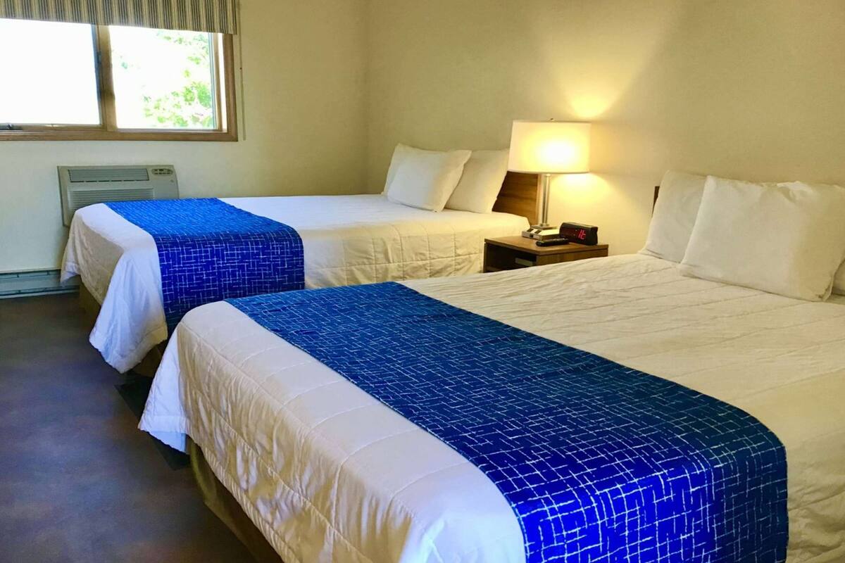A hotel room with two beds, each with a blue and white bedspread. There is a nightstand with a lamp and a phone, and a window with blinds.