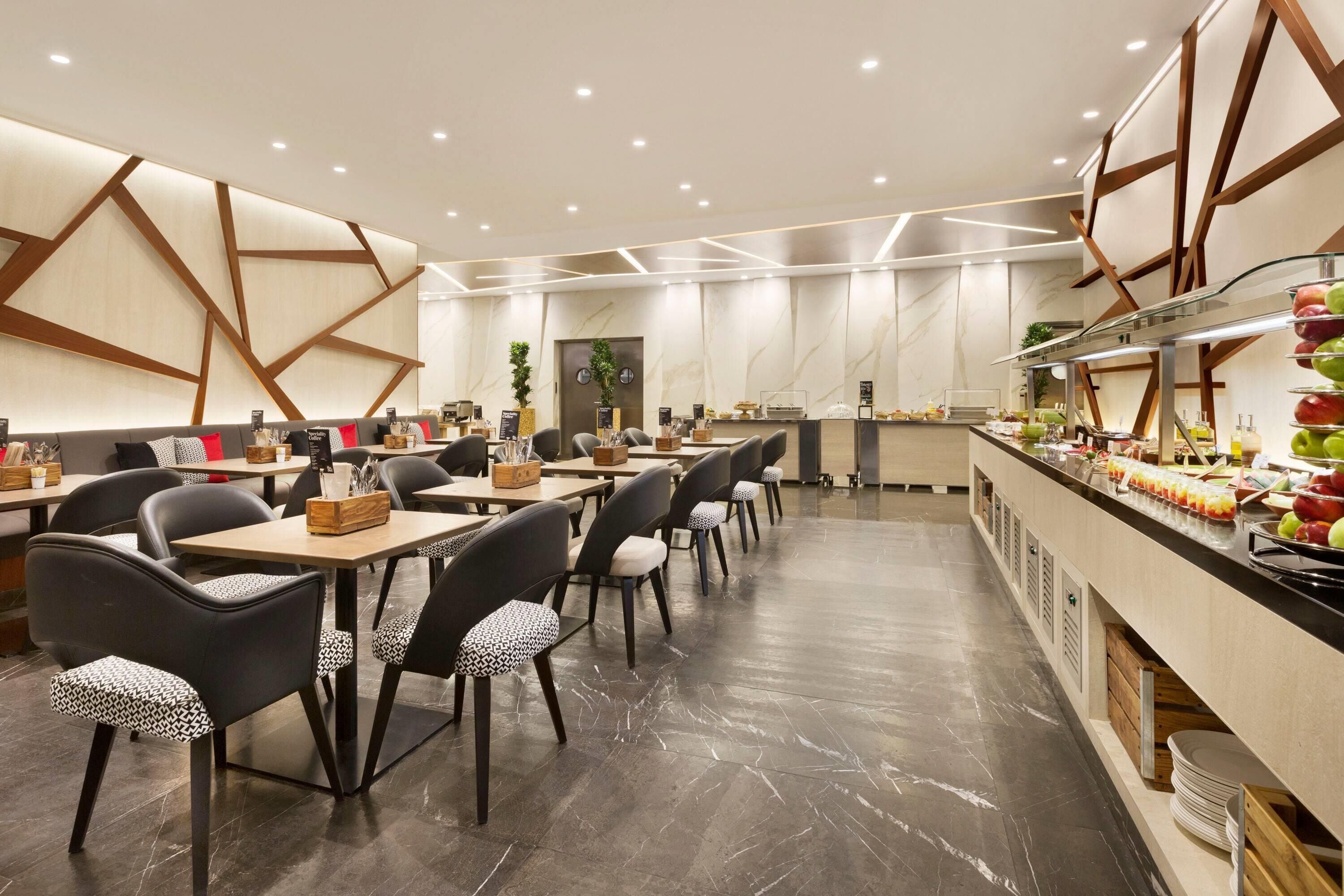 A modern dining area with marble flooring, black and patterned chairs, and a buffet counter with fruits and beverages.