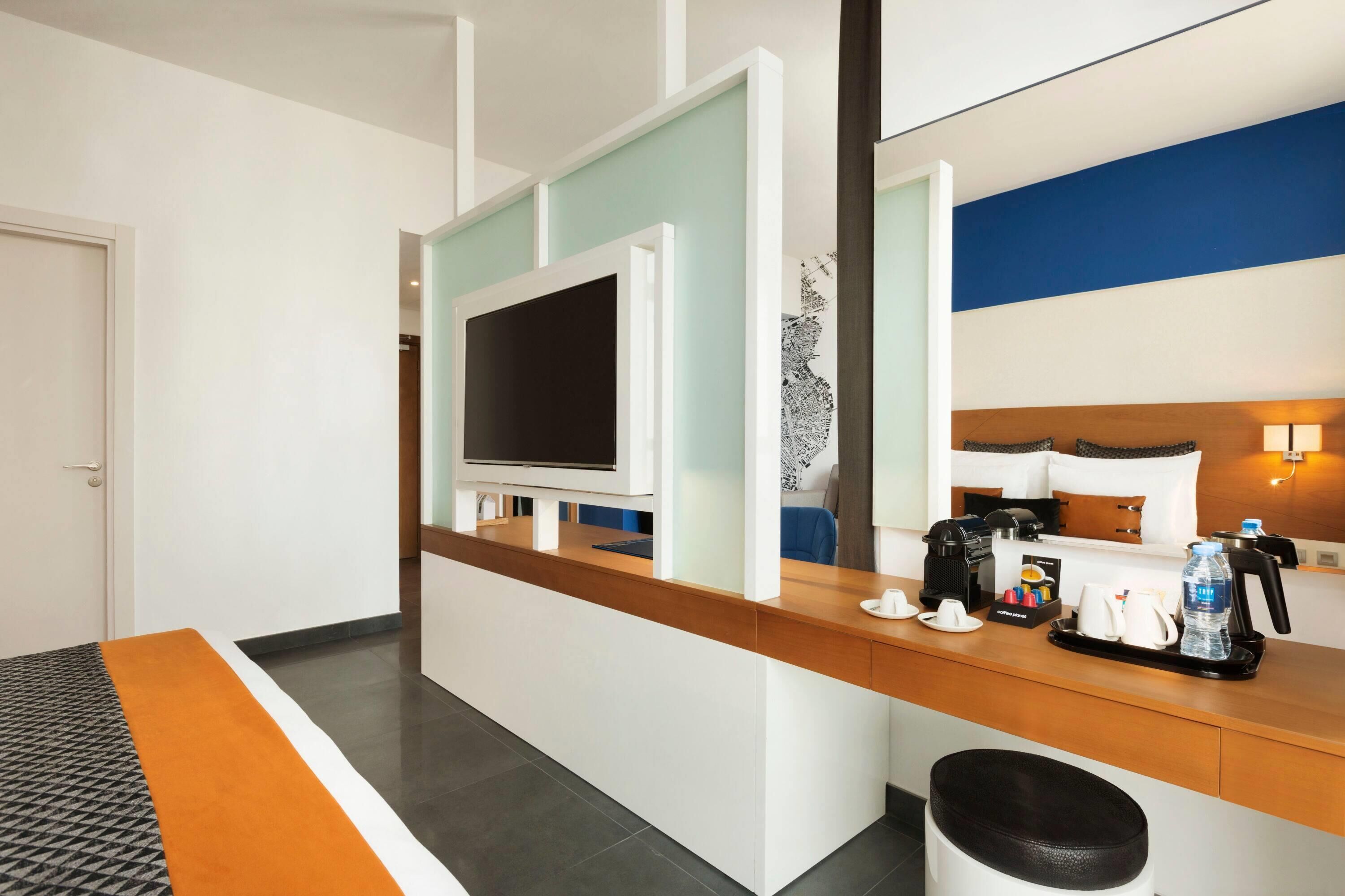 Habitació d'hotel amb taulell de recepció de fusta, televisió de pantalla plana, llit amb capçalera, cafetera i safata de recollida de basures.