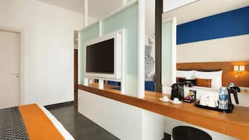 A hotel room with a wooden reception desk, a flat-screen TV, a bed with a headboard, a coffee maker, and a trash bin.