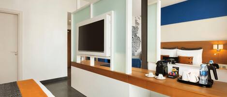 A hotel room with a wooden reception desk, a flat-screen TV, a bed with a headboard, a coffee maker, and a trash bin.