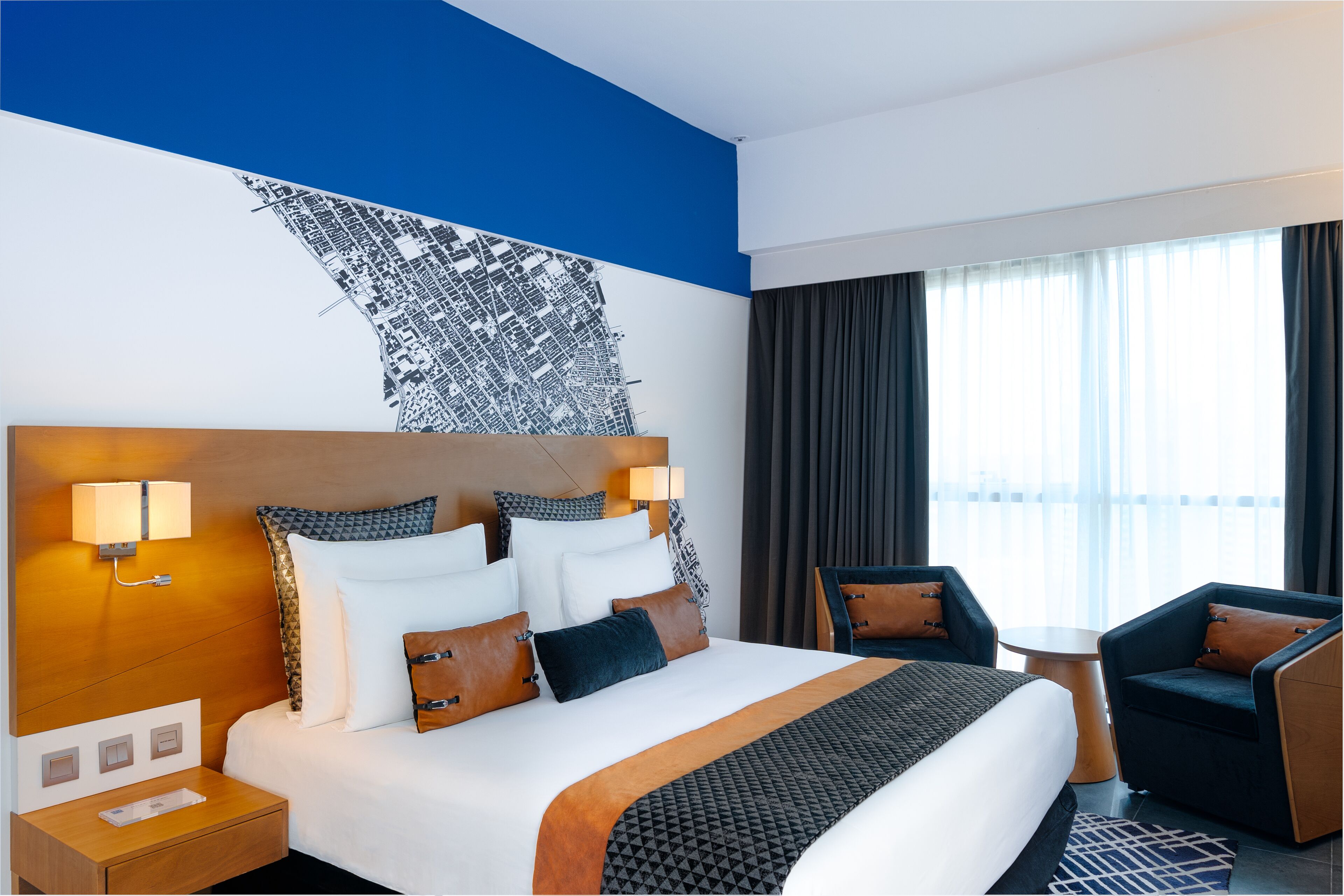 Ett hotellrum med en stor säng, två sänglampor, en sänggavel med en målning som föreställer en stadsbild, ett litet bord, två fåtöljer och utsikt över staden genom stora fönster.
