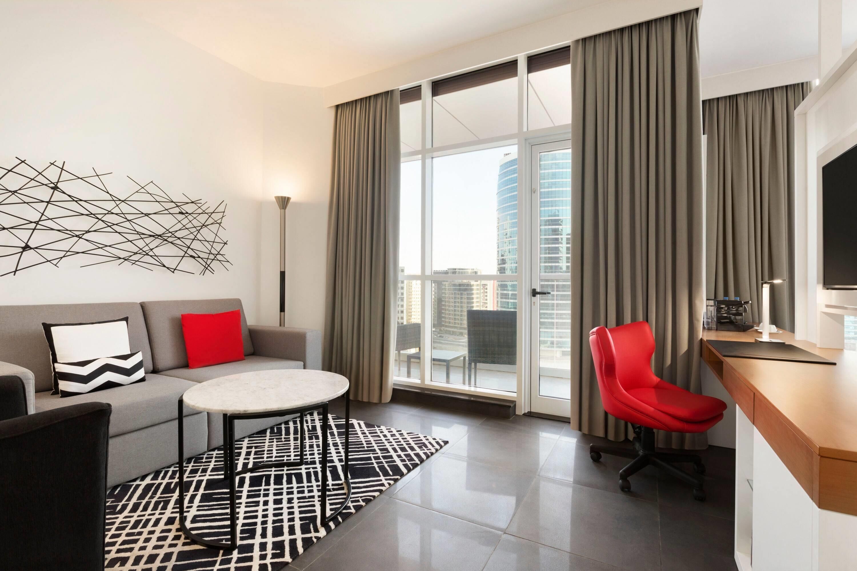 A modern hotel room with a sofa, a round table, a red office chair, a TV, and a large window with curtains.
