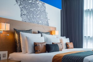 A modern hotel room with a large bed, multiple pillows, a headboard with a cityscape mural, and two wall-mounted lamps.