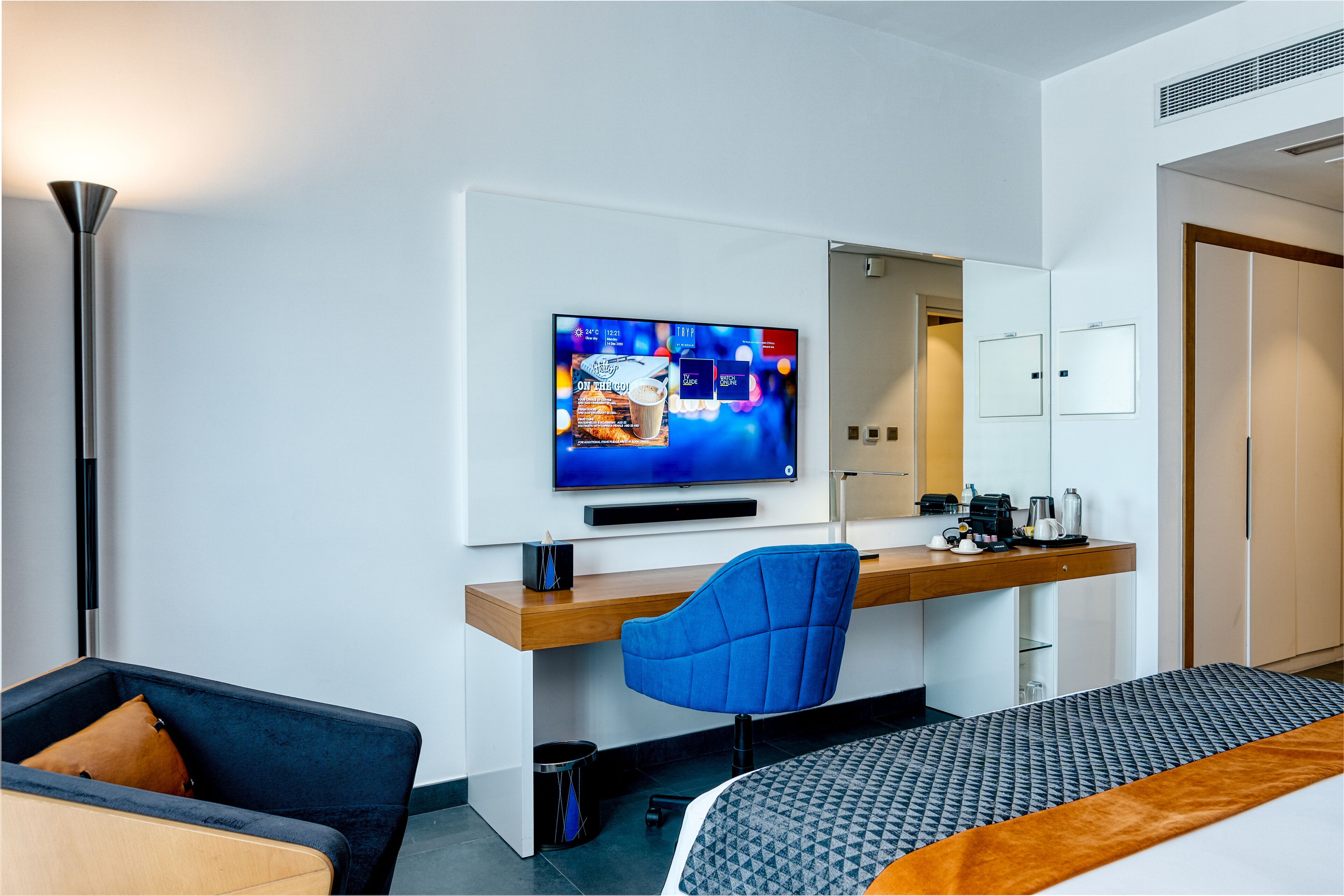 A modern hotel room with a flat-screen TV mounted on the wall, a wooden desk with a chair, a bed with a patterned bedspread, and a large mirror above the desk.