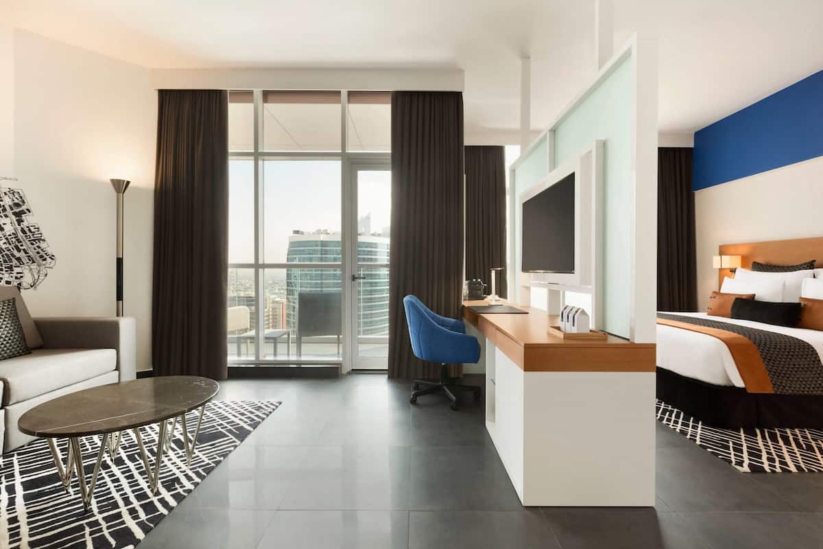 A modern hotel room with a large window, a desk, a chair, a bed, and a television.
