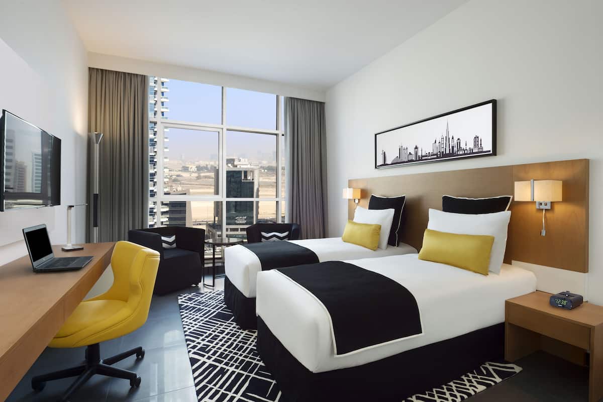 A hotel room with two beds, a desk with a laptop, a chair, a TV, and a city view.