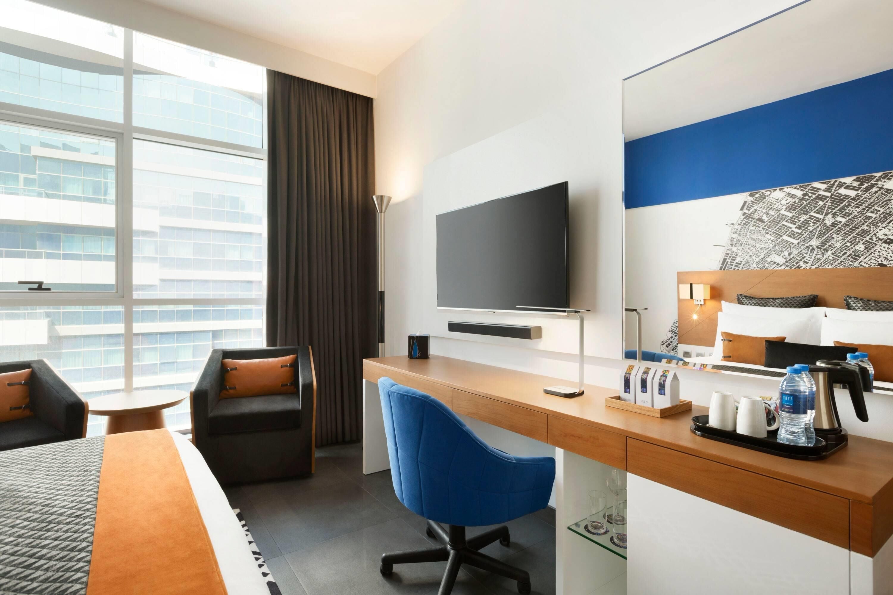 A hotel room with a bed, a desk with a computer, a TV, a chair, a coffee maker, and a window with a view of buildings.