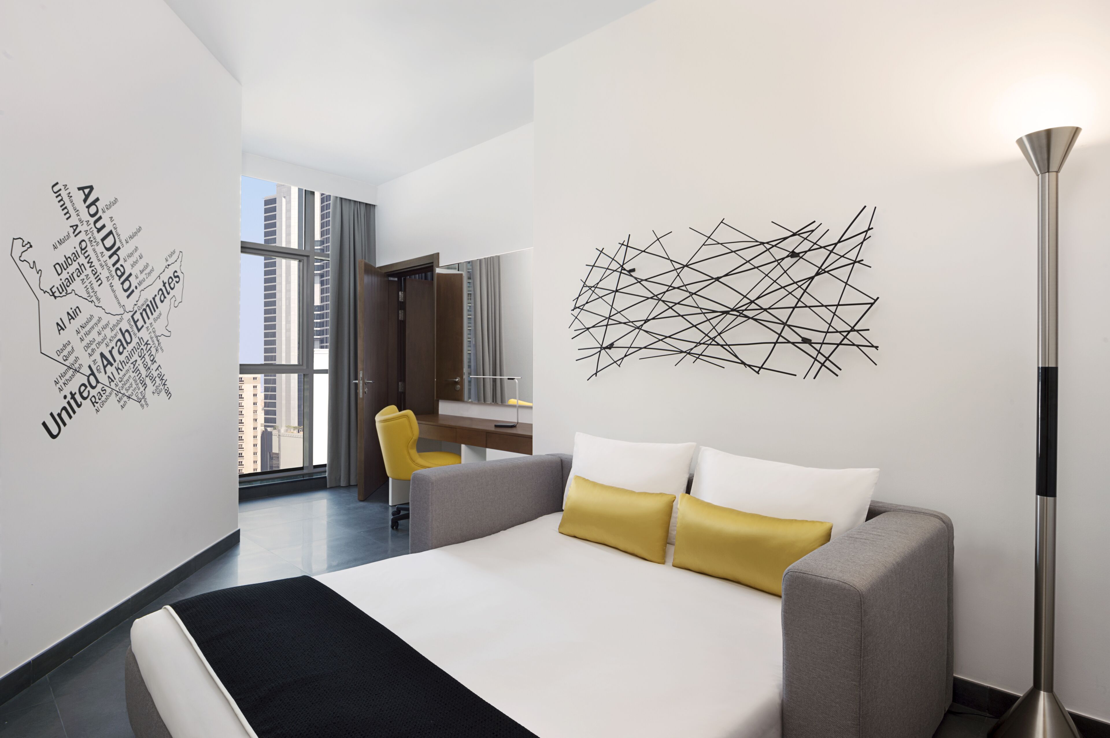 A modern hotel room with a bed, a desk, a chair, and a view of the cityscape.