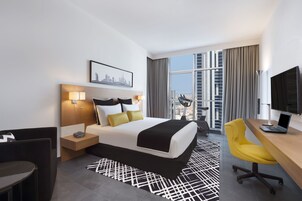 Habitación de hotel con una cama grande, un escritorio con una computadora portátil, una silla, un televisor y vistas a la ciudad.