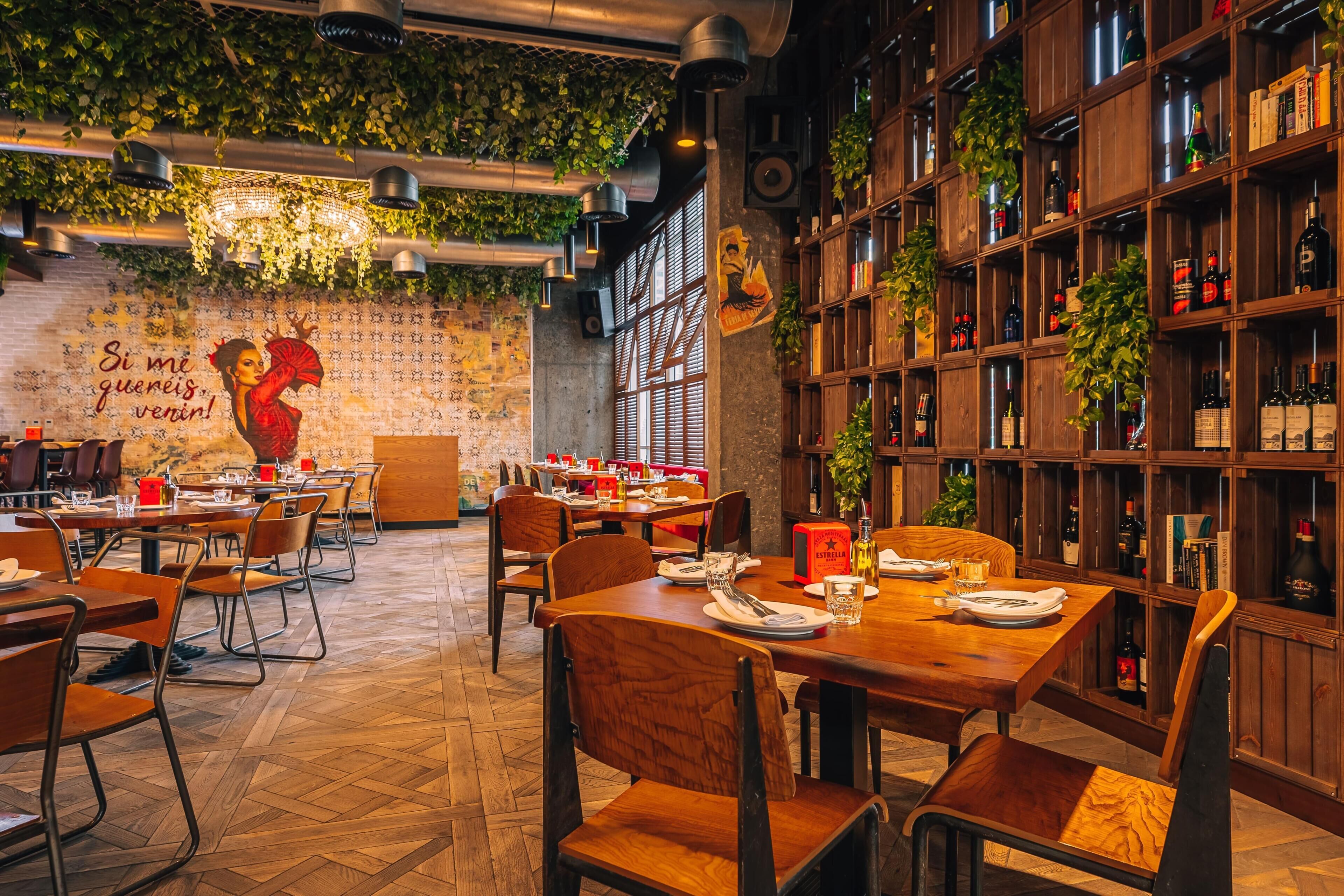 A restaurant with wooden tables and chairs, a wall mural, and shelves with bottles.