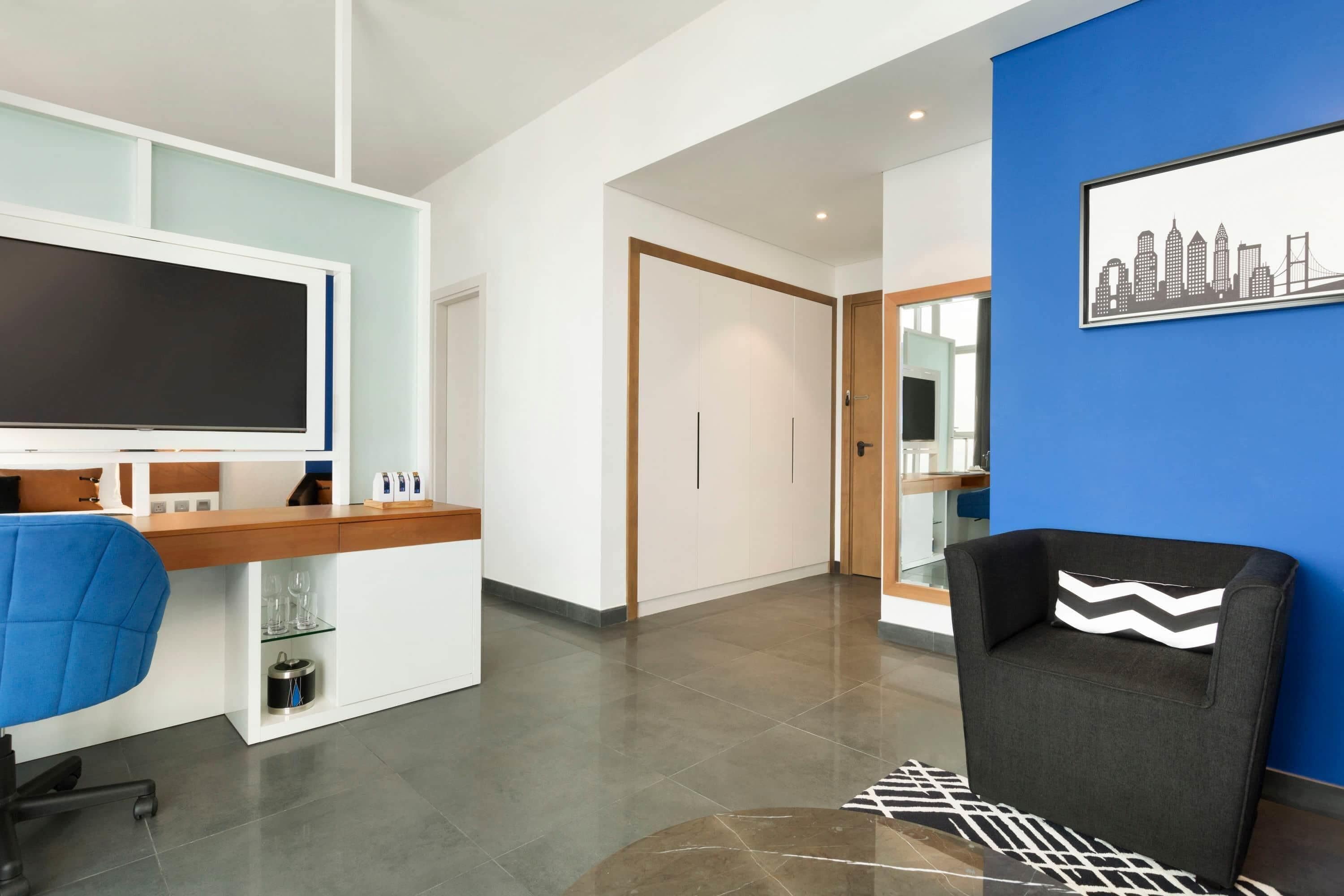 A modern hotel lobby with a reception desk, a blue wall with a cityscape mural, a black armchair, and a patterned rug.