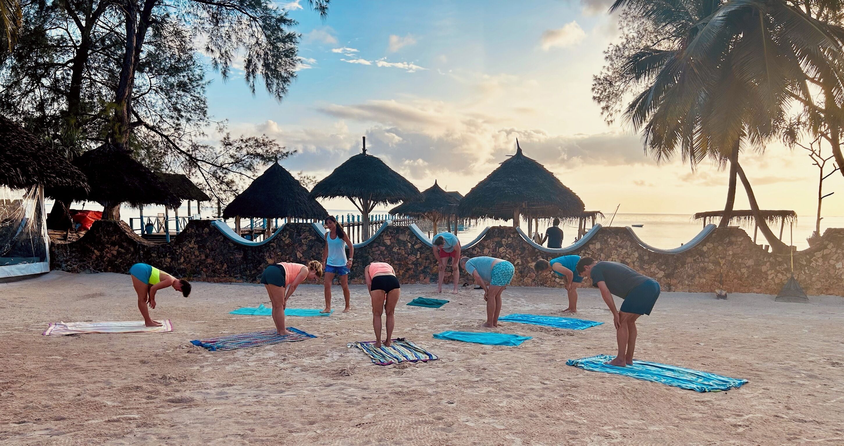 Une plage où des personnes pratiquent le yoga sur des tapis, entourées de constructions à toits de chaume et de palmiers.