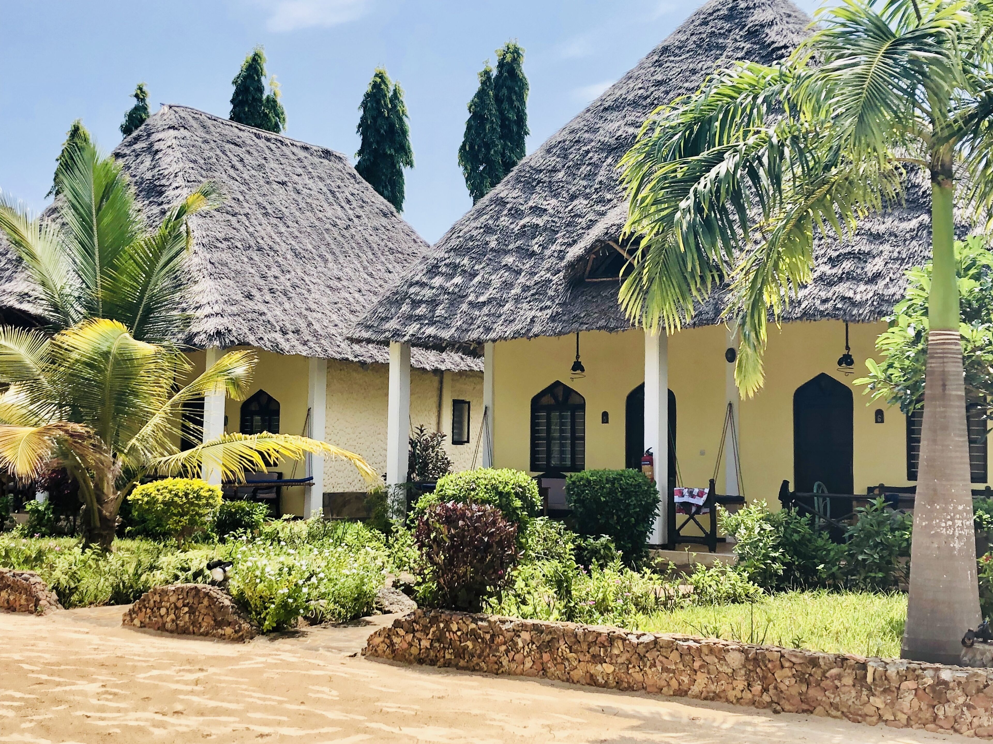 A row of thatched-roof huts with a yellow exterior, surrounded by greenery and a stone wall.