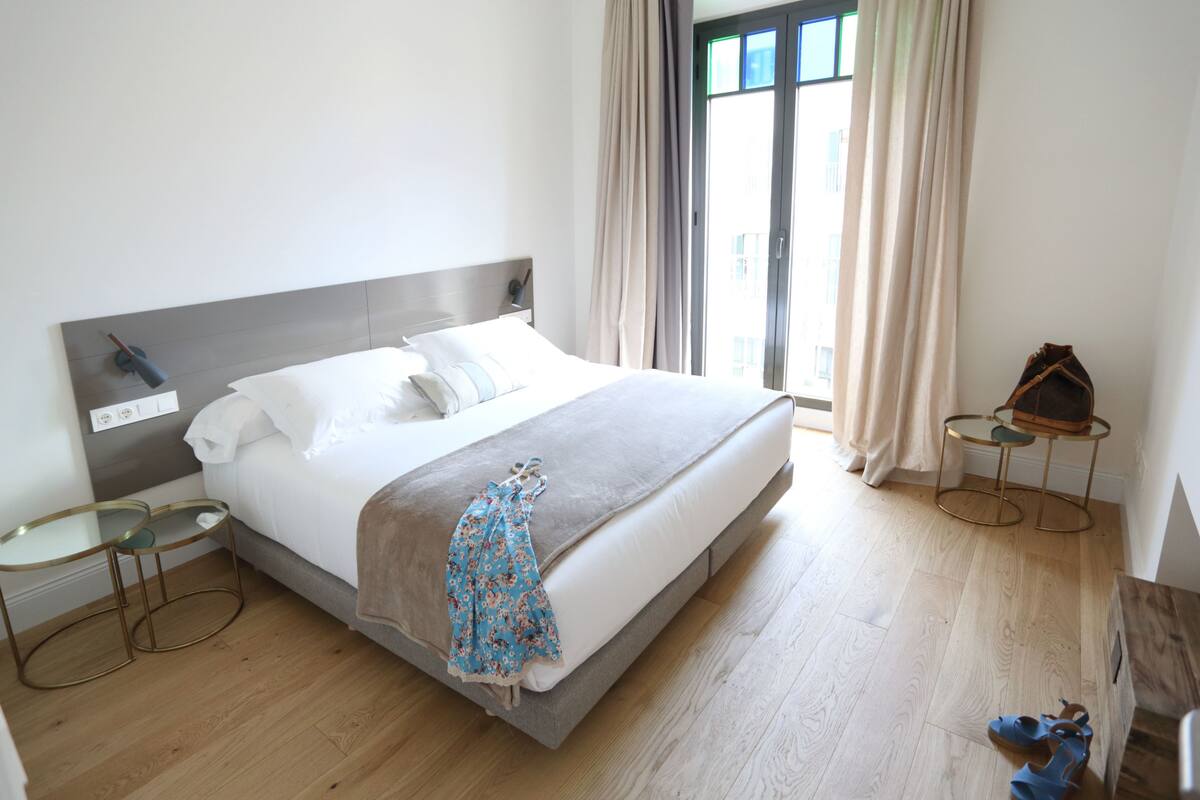 A modern hotel room with a large bed, a bedside table, a small round table, and a view of the outside through a window.