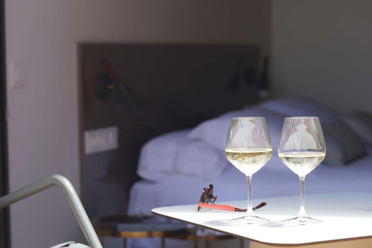 Two glasses of white wine on a white table in a hotel room with a bed in the background.